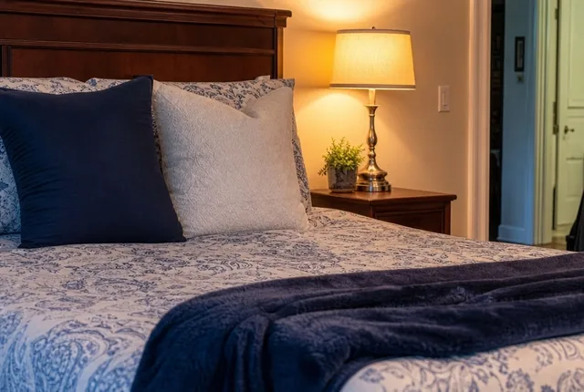 Pillows and a folded blanket on a made-up bed beside a lit bedside lamp