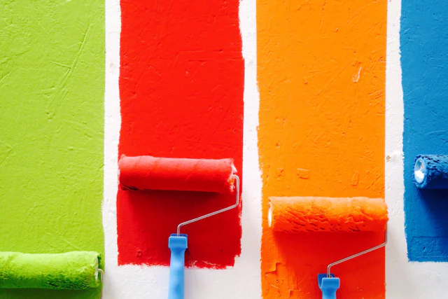 Four paint rollers leaving green, red, orange and blue stripes on a white wall